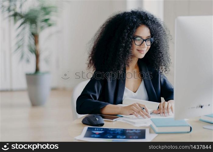 Freelance Afro woman works remotely, writes information, focused at computer screen with delighted expression, poses at workplace in spacious cabinet, potted plant in background. E learning concept