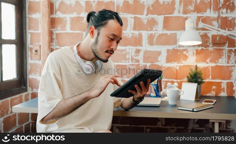 Freelance Asia guy using tablet for searching data and social media on internet in living room at house. Working from home, remotely work, social distancing, quarantine for coronavirus prevention.