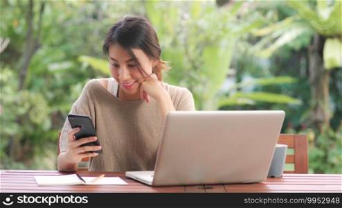 Freelance Asian woman working at home, business female working on laptop and using mobile phone drinking coffee sitting on table in the garden in morning. Lifestyle women working at home concept.