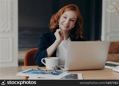 Freelance business woman with ginger hair sits in front of laptop computer, communicates with colleagues via video conference, sits at desktop, drinks coffee, has happy expression. Distance job