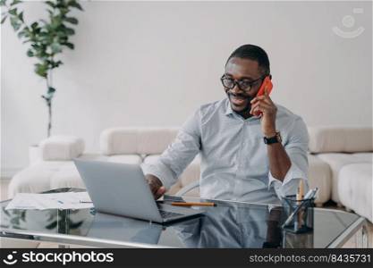 Freelancer is working online and having conversation with client on phone. Happy afro businessman has online meeting. Workplace at home office. Remote african american worker.. Freelancer is working online and having conversation or meeting with client on phone.
