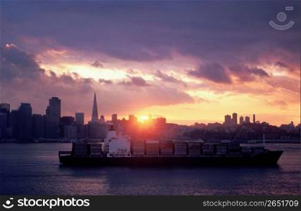Freighter of containers with cityscape and sunset in background