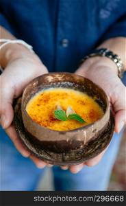 French dessert, Hands holding beautiful creamy vanilla Creme Brulee in coconut shell. Close up shot from front