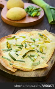 French vegetarian Tarte Flambee made of thin bread dough with a cream and leek topping, served on wooden board with ingredients in the back (Selective Focus, Focus on the upper part of the first piece)