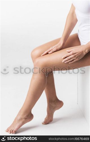 Fresh and beautiful. Attractive young woman in tank top and panties sitting on cube and touching legs against white background. Woman touching legs