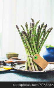 Fresh asparagus bunch with blank label on kitchen table at window background, front view. Healthy seasonal food concept