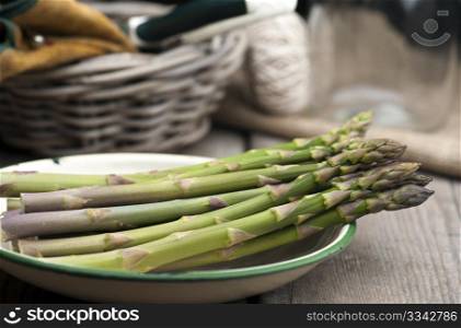 Fresh Asparagus Laid Out In An Enamel Dish On A Rustic Kitchen Table, With A Basket, Gardening Gloves, Secateurs and String In The Background