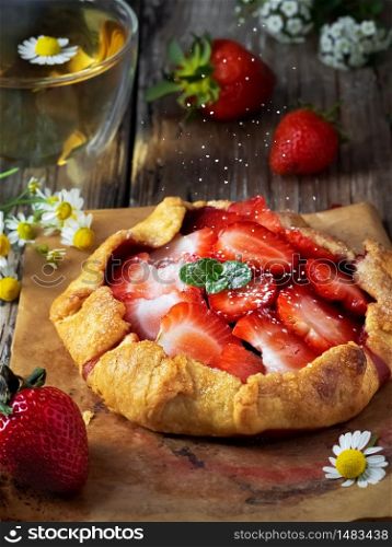 Fresh baked galette pie or open strawberry pie on a wooden table, closeup. Galette is sprinkled with powdered sugar. Homemade cakes, fresh berries and a cup of chamomile tea