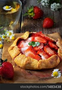 Fresh baked galette pie or open strawberry pie on a wooden table, closeup. Galette is sprinkled with powdered sugar. Homemade cakes, fresh berries and a cup of chamomile tea