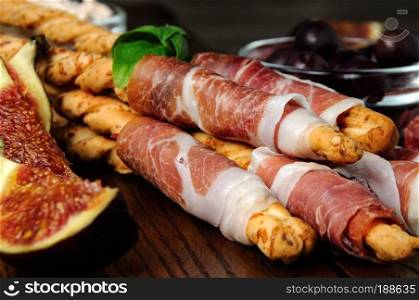  Fresh baked Italian breadsticks with Parmesan wrapped with a slice of prosciutto and olives, figs. Italian dish with antipasto on a wooden table.