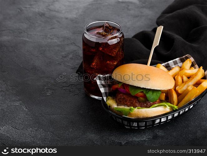 Fresh beef burger with sauce and vegetables and glass of cola soft drink with potato chips fries in black serving basket on stone kitchen background.