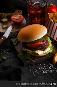 Fresh beef burger with sauce and vegetables and glass of cola soft drink with potato chips fries on stone kitchen background.