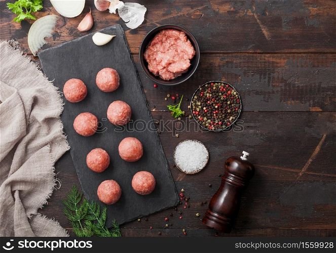 Fresh beef raw meatballs on stone board with mince on bowl plate with pepper, salt and garlic on dark board with parsley and dill with onion. Space for text