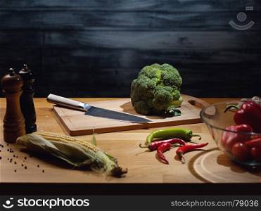 Fresh broccoli on wooden board with knife. Low key shot, light on board, some vegetables around on table. Copy space.