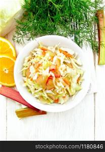 Fresh cabbage, carrot and rhubarb salad with orange juice, honey and mayonnaise dressing in a plate on a towel, dill and fork on wooden board background from above