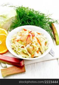 Fresh cabbage, carrot and rhubarb salad with orange juice, honey and mayonnaise dressing in a plate on a towel, dill and fork on white wooden board background 