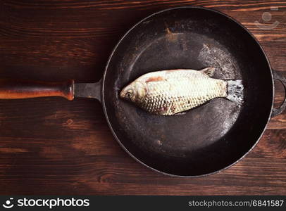 Fresh carp fish in a black cast-iron frying pan on a brown wooden table, top view