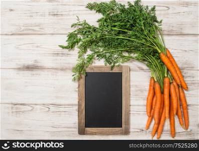 Fresh carrots roots with green leaves and blackboard over wooden background. Organic food concept