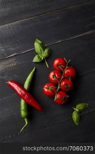 Fresh cherry tomatoes on a branch, green and red chilies and basil leaves on a dark wooden background with copy space. Organic vegetables. Flat lay. A composition of fresh vegetables, tomatoes, chili and basil leaves on a dark wooden background. Flat lay