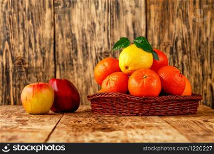 Fresh citrus fruits, tangerines and lemon with green leaves in wicker basket, red ripe apples lie on an old wooden table on wooden background, image with copy space.. Tangerines and lemon in a wicker basket, red apples lie on an old wooden table.