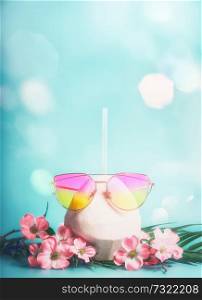 Fresh coconut cocktail with pink sunglasses with tropical leaves and flowers at blue background with bokeh. Tropical vacation. Summer holiday. Beach party