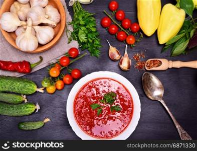 Fresh cold tomato and vegetable soup gazpacho in a white round plate with an iron spoon on a black background, around the ingredients and spices