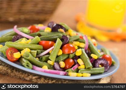 Fresh colorful vegetarian salad made of green beans, cherry tomatoes, sweet corn, black olives and red onions on blue plate with orange juice and basket in the back (Selective Focus, Focus one third into the salad)