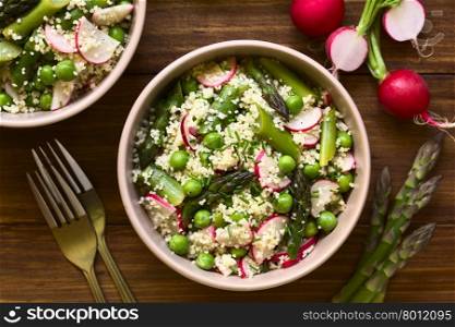 Fresh couscous salad with green asparagus, peas, radish slices and chives, photographed overhead on dark wood with natural light (Selective Focus, Focus on the top of the salad)