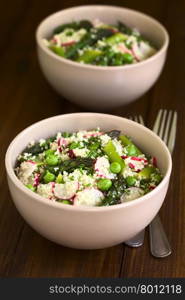 Fresh couscous salad with green asparagus, peas, radish slices and chives served in bowls, photographed on dark wood with natural light (Selective Focus, Focus in the middle of the first salad)