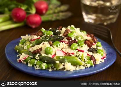 Fresh couscous salad with green asparagus, peas, radish slices and fried bacon pieces served on plate, photographed on dark wood with natural light (Selective Focus, Focus in the middle of the salad)