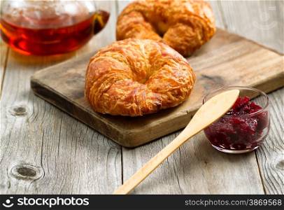Fresh croissants, focus on front of first croissant, with fresh cranberry jam and tea for breakfast. Close up image with rustic wood underneath.