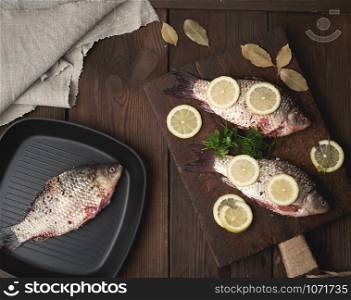 fresh crucian fish sprinkled with spices and lemon slices and lies on a brown wooden cutting board, wooden table from boards, top view