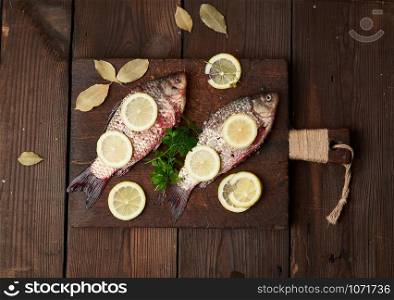 fresh crucian fish sprinkled with spices and lemon slices and lies on a brown wooden cutting board, wooden table from boards, top view