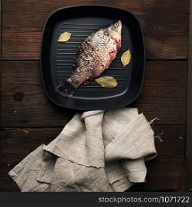 fresh crucian fish sprinkled with spices and lies in a black square pan, wooden table from boards, top view