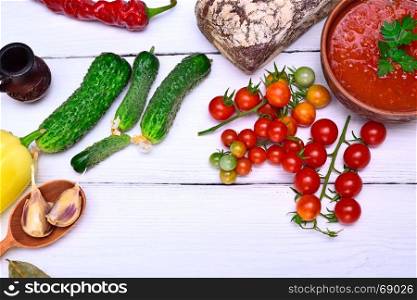 Fresh cucumber and red cherry tomatoes and a plate of cold gazpacho soup on a white table, top view, empty space in the middle