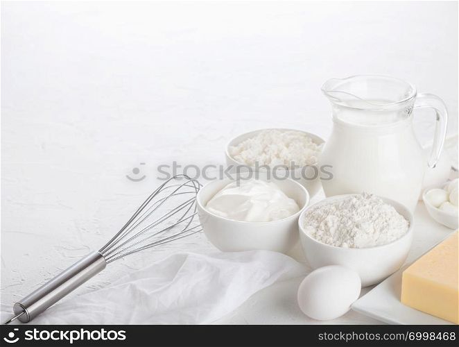 Fresh dairy products on white table background. Glass jar of milk, bowl of sour cream, cottage cheese and baking flour and mozzarella. Eggs and cheese. Steel whisk