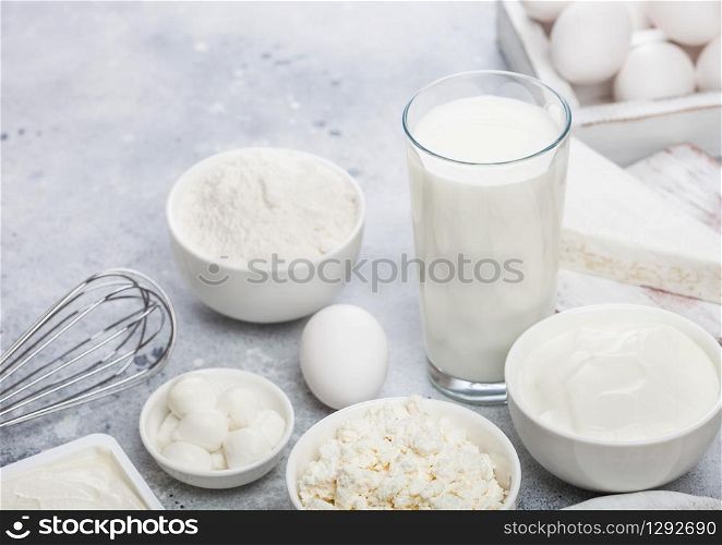 Fresh dairy products on white table background. Glass of milk, bowl of sour cream, cottage cheese and baking flour and mozzarella. Eggs and cheese. Steel whisk