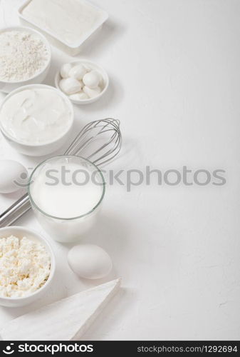 Fresh dairy products on white table background. Glass of milk, bowl of flour, sour cream and cottage cheese and eggs. Steel whisk