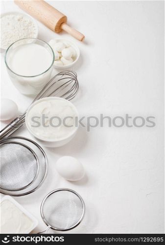 Fresh dairy products on white table background. Glass of milk, bowl of flour, sour cream and cottage cheese and eggs. Steel whisk and rolling pin.