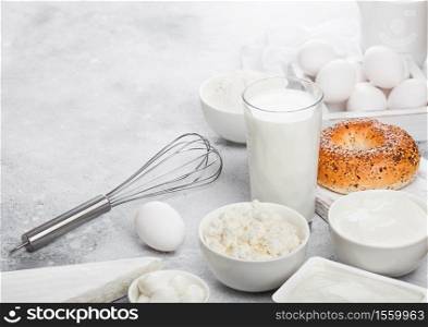 Fresh dairy products on white table background. Glass of milk, bowl of sour cream and cottage cheese and eggs in wooden box. Fresh baked baggel on round chopping board. Steel whisk.