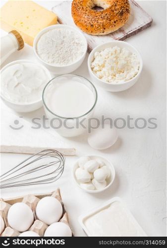 Fresh dairy products on white table background. Glass of milk, bowl of flour and cottage cheese and eggs. Fresh baked bagel with knife. Steel whisk.