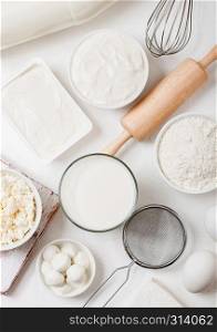Fresh dairy products on white table background. Glass of milk, bowl of flour, sour cream and cottage cheese and eggs. Steel whisk and rolling pin.