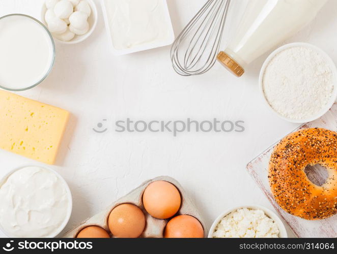 Fresh dairy products on white table background. Glass of milk, bowl of sour cream and cottage cheese and eggs. Fresh baked bagel. Steel whisk. Top view.