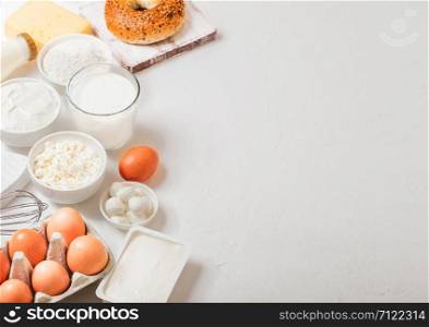 Fresh dairy products on white table background. Glass of milk, bowl of sour cream and cottage cheese and eggs. Fresh baked bagel. Steel whisk. Top view.