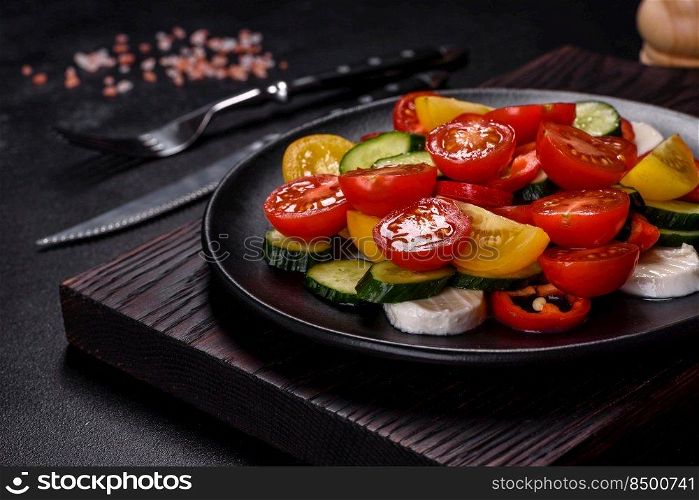 Fresh, delicious salad with cherry tomatoes, cucumbers, sweet peppers, cheese and olive oil on a black plate against a dark concrete background. Vegetarian dish. Fresh, delicious salad with cherry tomatoes, cucumbers, sweet peppers, cheese and olive oil