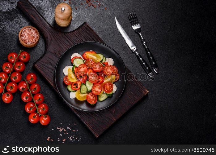 Fresh, delicious salad with cherry tomatoes, cucumbers, sweet peppers, cheese and olive oil on a black plate against a dark concrete background. Vegetarian dish. Fresh, delicious salad with cherry tomatoes, cucumbers, sweet peppers, cheese and olive oil