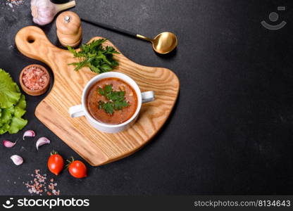 Fresh delicious tomato soup with spices and parsley in a white bowl on a wooden board against a dark concrete background. Fresh delicious tomato soup with spices and parsley in a white bowl on a wooden board