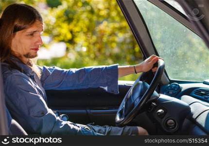 Fresh driver, traveling concept. Young man wearing jeans shirt having long hair driving car carefully on sunny day. Young man with long hair driving car