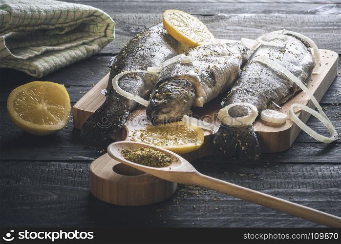 Fresh fish, placed on a wooden cutting board and sprinkled with herbs, slices of lemon and onion, on a rustic black table, under natural light.