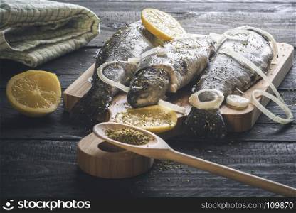 Fresh fish, placed on a wooden cutting board and sprinkled with herbs, slices of lemon and onion, on a rustic black table, under natural light.